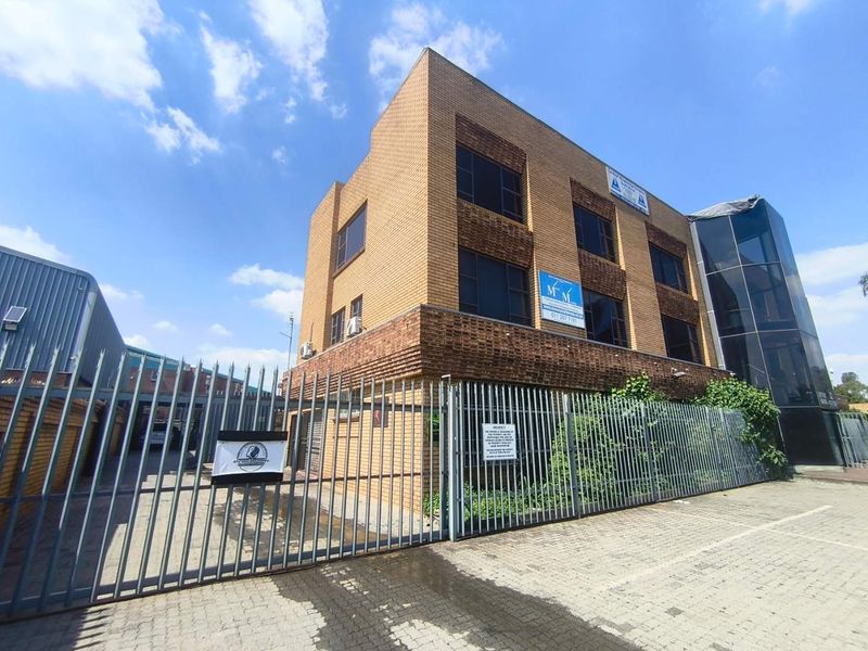 Front exterior view of a multi-story brick office building featuring a secure gated entrance, paved driveway, and a modern glass facade section.