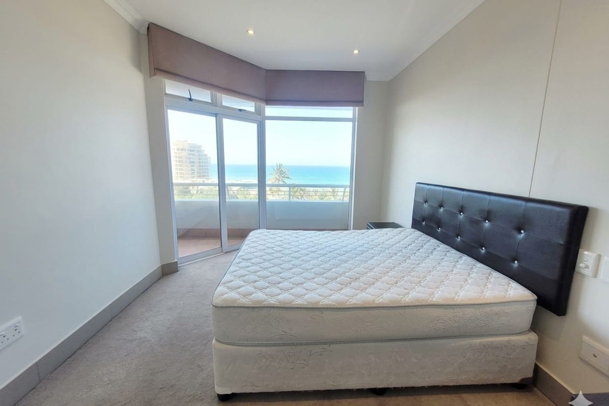 A bright bedroom featuring neutral walls, carpeted flooring, and a sliding glass door providing access to a balcony with ocean views.