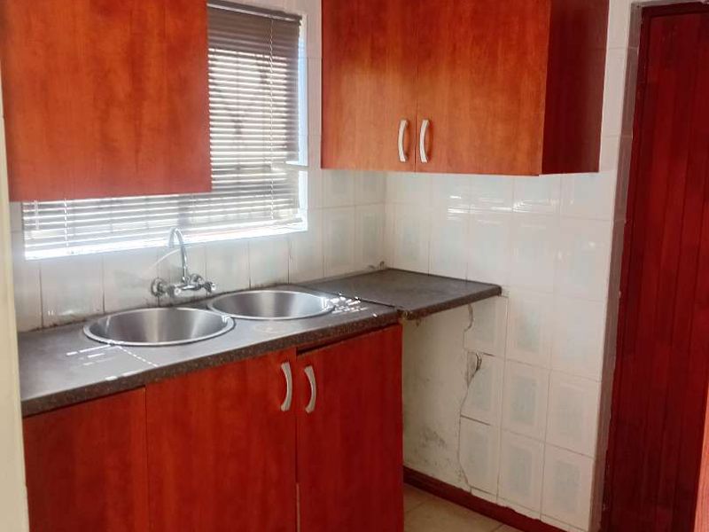 A compact kitchen featuring wooden cabinetry, a double bowl sink, tiled splashbacks, and tiled flooring, requiring some maintenance to the wall tiles and cabinetry.