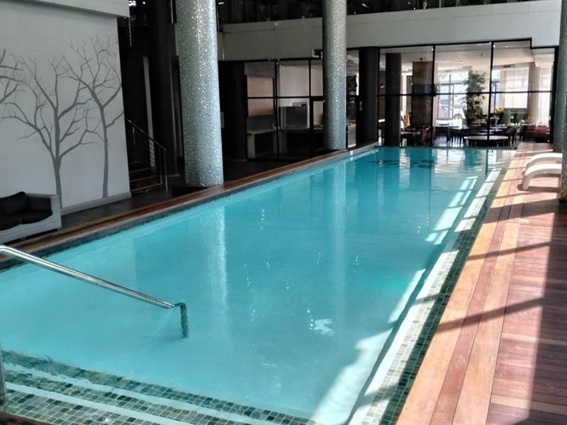 A modern indoor swimming pool area featuring timber decking, mosaic-tiled columns, and a mezzanine level, creating a sophisticated and well-maintained recreational space.