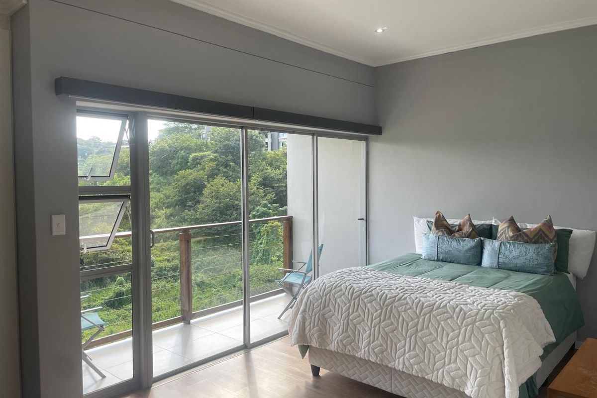Spacious bedroom featuring modern laminate flooring, neutral grey walls, and large sliding glass doors opening onto a private balcony with lush green views.