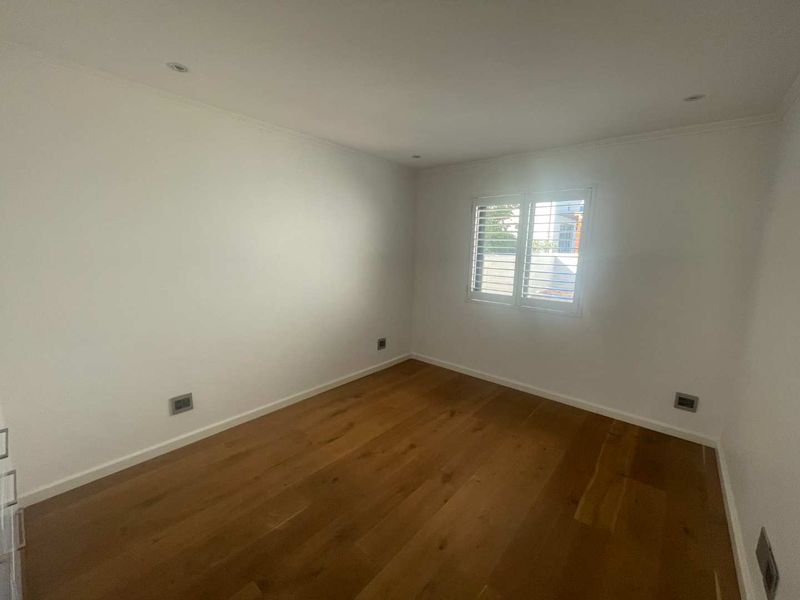 A bright, empty bedroom featuring polished hardwood flooring, recessed lighting, and white plantation shutters, offering a clean and neutral space.