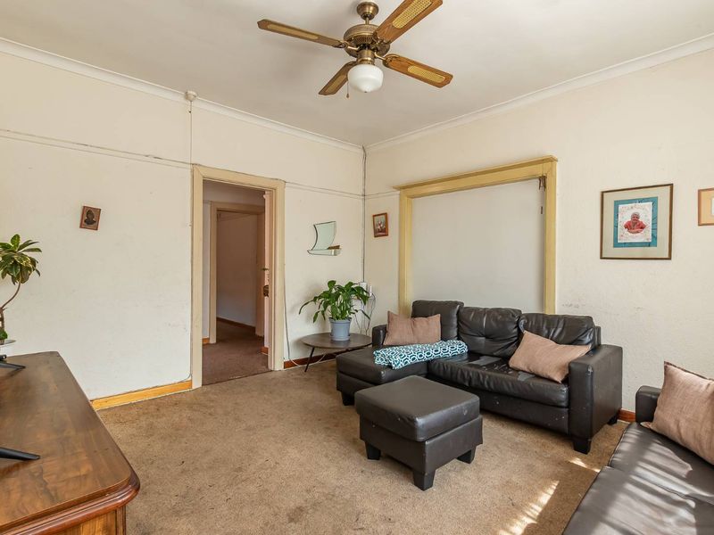 A living room featuring a ceiling fan, carpeted flooring, and traditional door architraves, currently requiring cosmetic updates and refurbishment to improve its overall condition.
