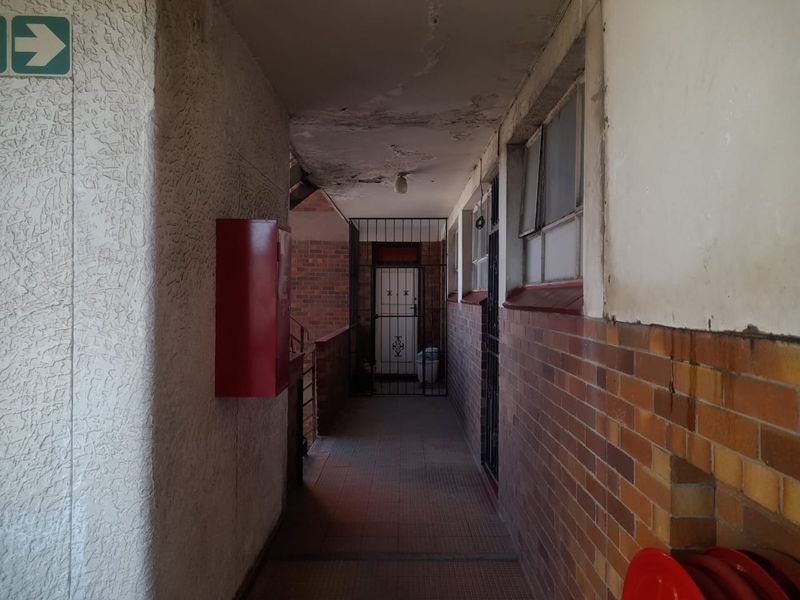 An exterior apartment corridor featuring brick walls, tiled flooring, security gates, and fire safety equipment, showing signs of ceiling wear and maintenance requirements.