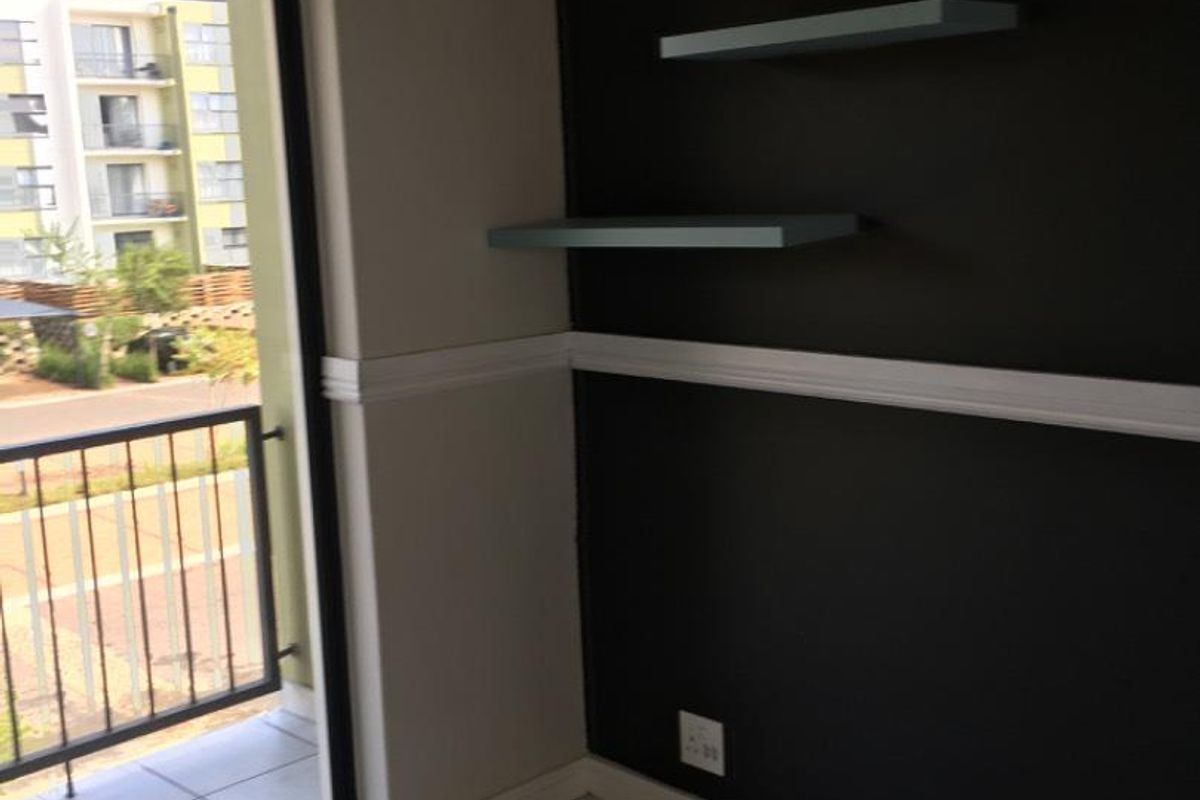 A bright living area featuring tiled flooring, a dark accent wall with built-in floating shelves, and direct access to a private balcony.