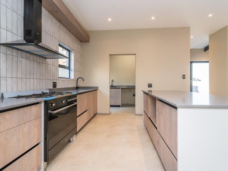 Modern kitchen featuring sleek wood-finish cabinetry, stone countertops, a gas stove with extractor fan, and a central island, with an adjacent scullery area.