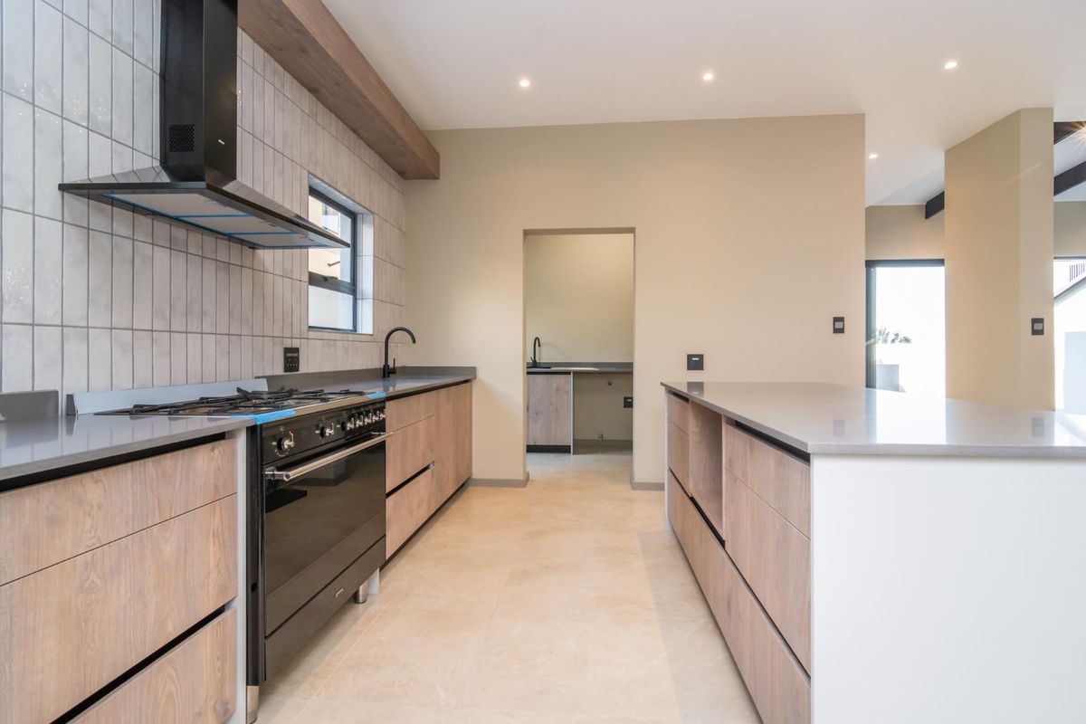 Modern kitchen featuring sleek wood-finish cabinetry, stone countertops, a gas stove with extractor fan, and a central island, with an adjacent scullery area.