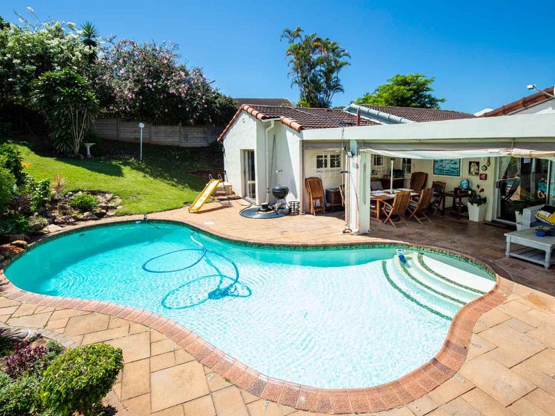A well-maintained outdoor entertainment area featuring a curved swimming pool, paved patio surround, and a covered patio space adjacent to the house.
