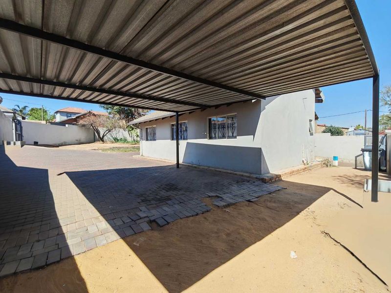 A covered carport area with a corrugated metal roof and paved driveway, featuring security bars on the exterior windows.