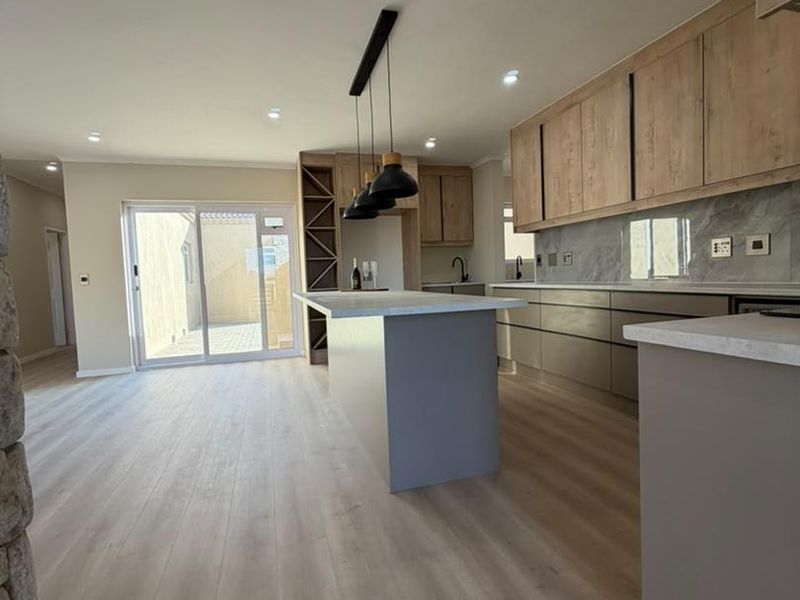 Modern open-plan kitchen and living area featuring light wood-look laminate flooring, stone countertops, sleek cabinetry, and direct access to an outdoor patio area.