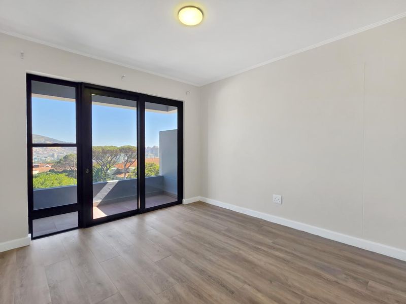 A bright, empty bedroom featuring modern laminate flooring, neutral walls, and a large sliding glass door providing access to a balcony with scenic views.