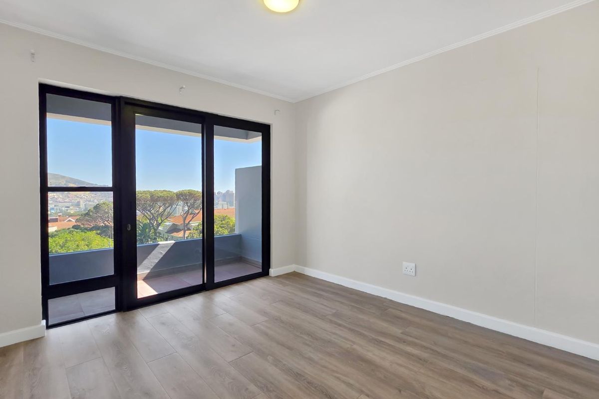 A bright, empty bedroom featuring modern laminate flooring, neutral walls, and a large sliding glass door providing access to a balcony with scenic views.
