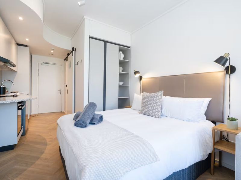 Modern open-plan studio apartment featuring a compact kitchen with stone countertops, herringbone flooring, and a built-in wardrobe with integrated shelving.