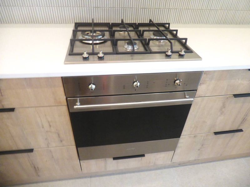 Modern kitchen featuring a stainless steel gas cooktop, built-in oven, stone countertops, and contemporary timber-look cabinetry with a tiled splashback.