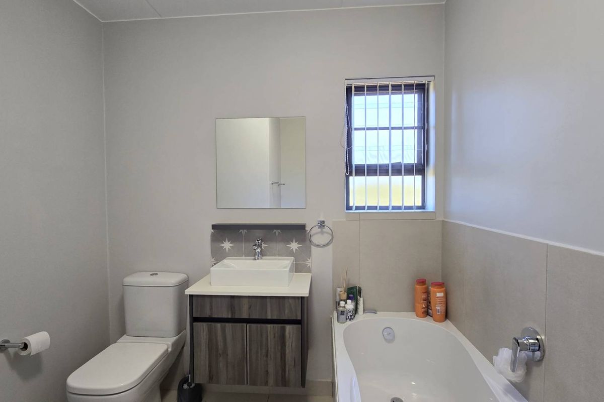 A clean and modern bathroom featuring a white bathtub, a contemporary vanity with a vessel sink, a wall-mounted mirror, and a window with security bars.