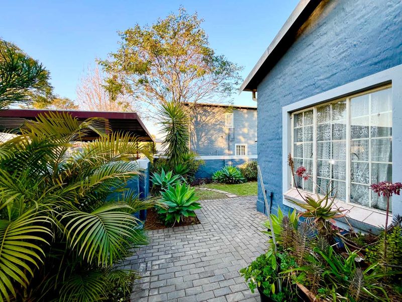 A well-maintained rear exterior area featuring a paved walkway, established garden landscaping, and a blue brick facade with secure window detailing.