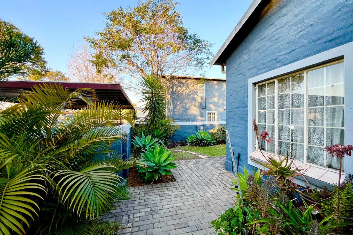 A well-maintained rear exterior area featuring a paved walkway, established garden landscaping, and a blue brick facade with secure window detailing.