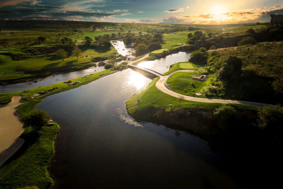 An aerial view of a scenic golf course landscape featuring a winding river, a pedestrian bridge, and lush green fairways at sunset.