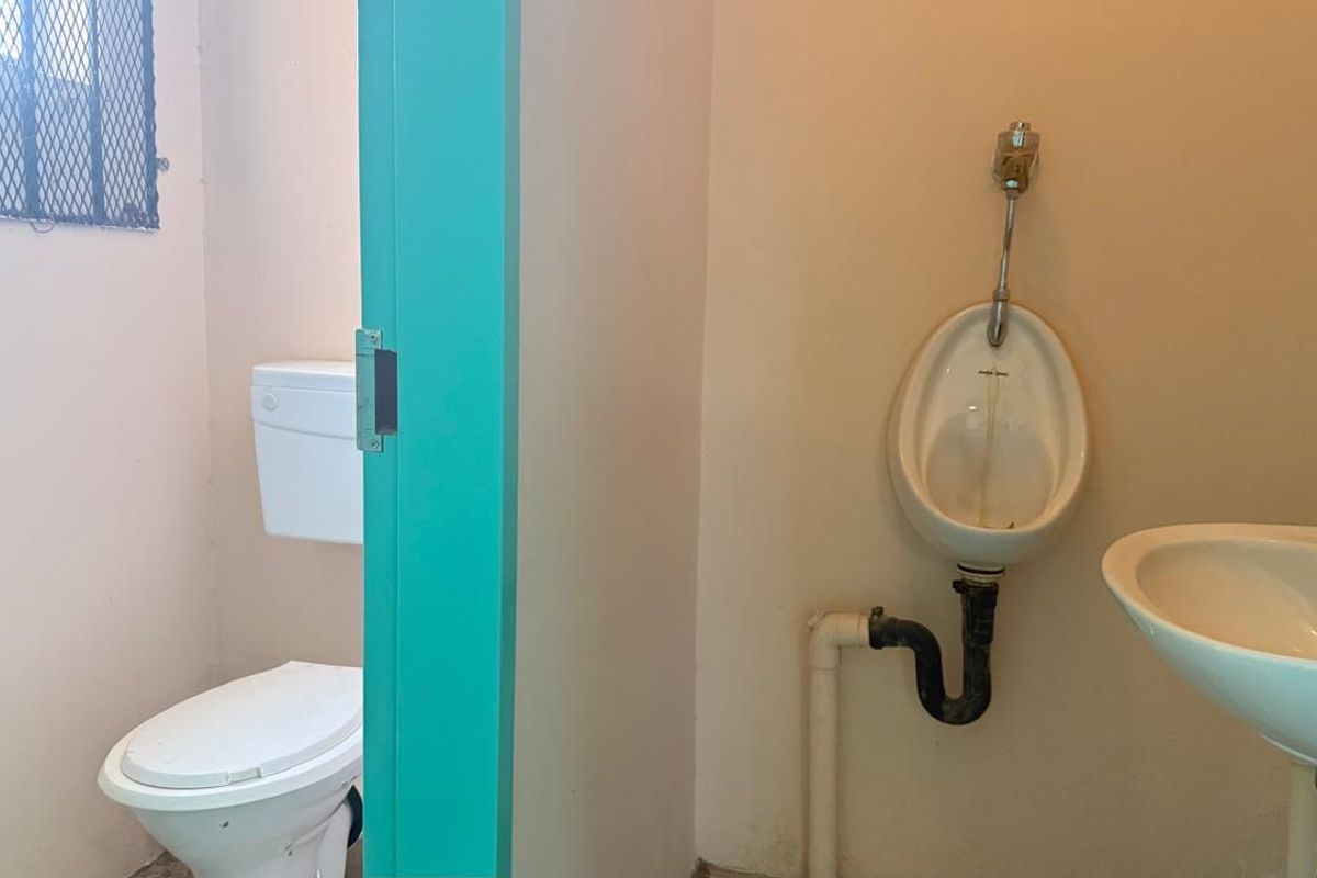 A functional bathroom facility featuring a wall-mounted urinal, pedestal basin, and standard toilet, currently in need of cosmetic updates and maintenance.