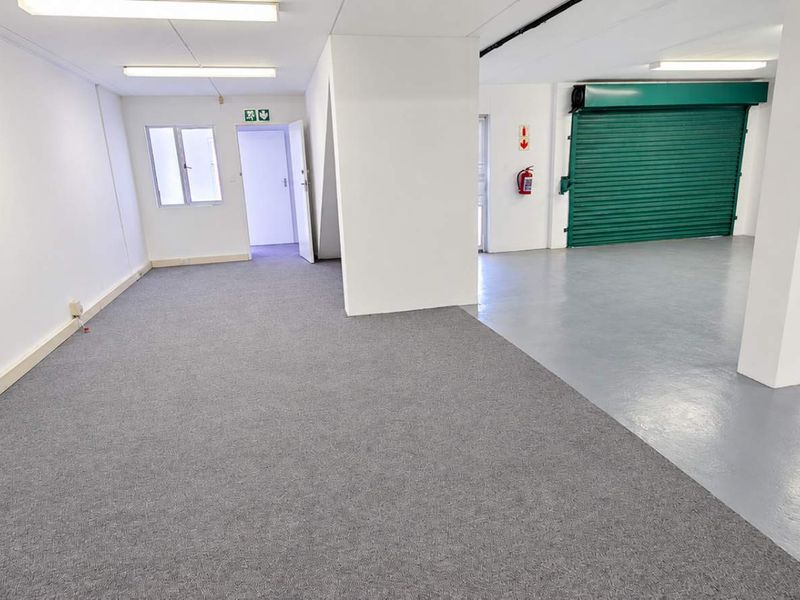 Spacious open-plan industrial unit featuring a combination of carpeted and epoxy-coated flooring, roller shutter access, and functional overhead lighting.