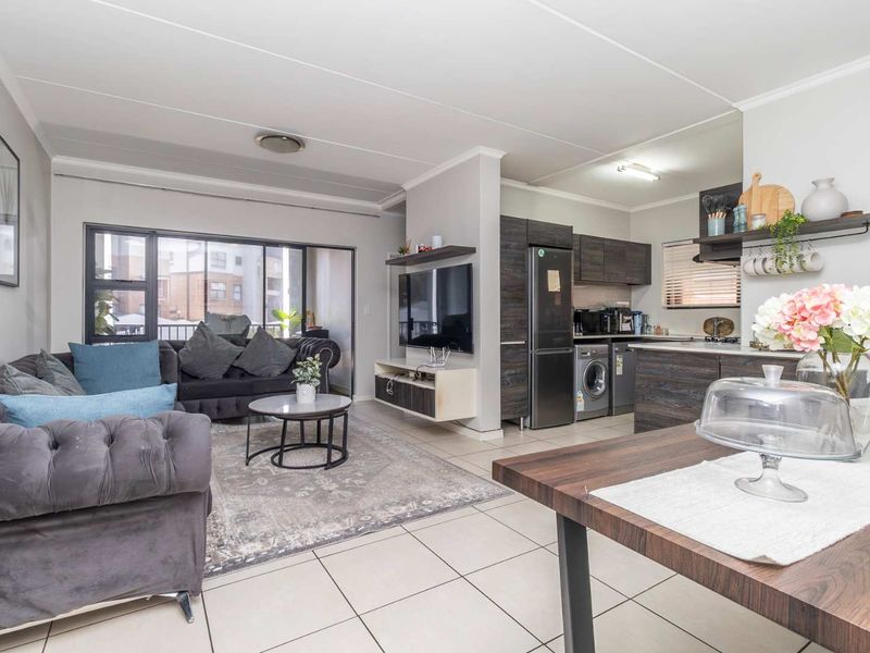 A bright open-plan living and kitchen area featuring tiled floors, modern dark wood cabinetry, and a seamless transition to a balcony through sliding glass doors.