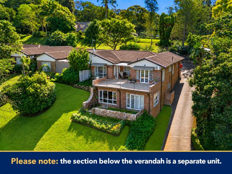 Aerial view of a brick townhouse featuring a spacious upper balcony, landscaped garden surrounds, and a private driveway.