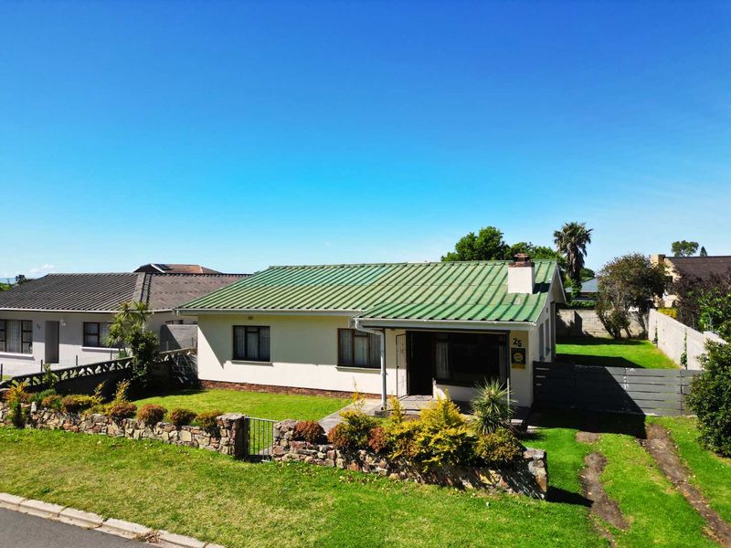 Front exterior view of a single-story house featuring a green corrugated metal roof, stone boundary wall, and a private driveway.
