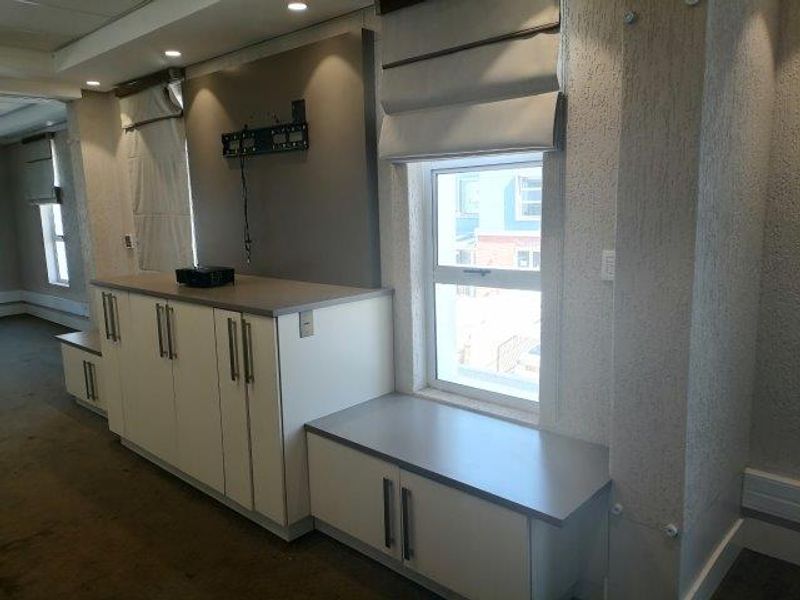 A professional office space featuring built-in white cabinetry, integrated storage, recessed ceiling lighting, and large windows with fitted roman blinds.