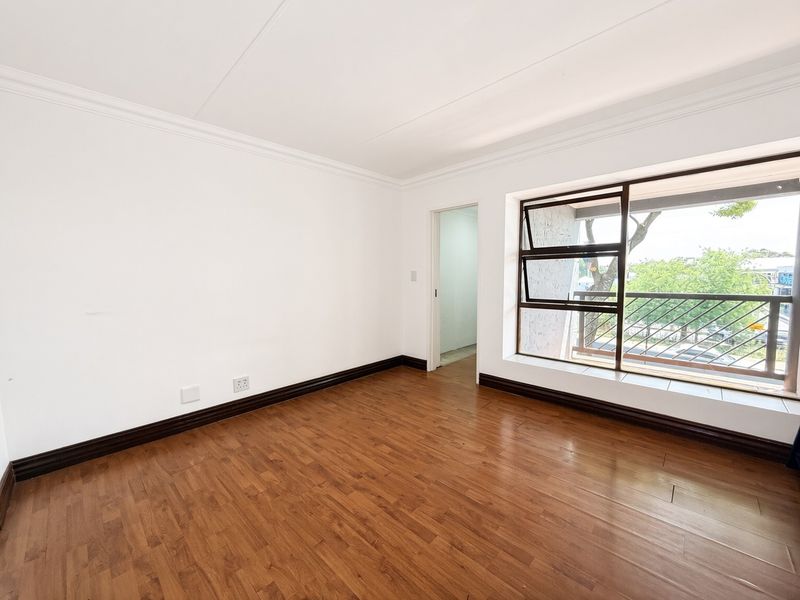 A bright, empty bedroom featuring laminate flooring, white walls with cornice detailing, and a large window with security bars overlooking the exterior.
