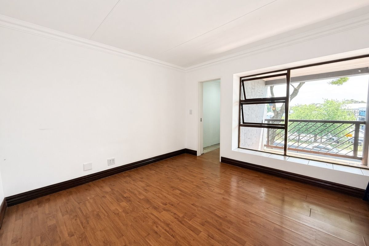A bright, empty bedroom featuring laminate flooring, white walls with cornice detailing, and a large window with security bars overlooking the exterior.