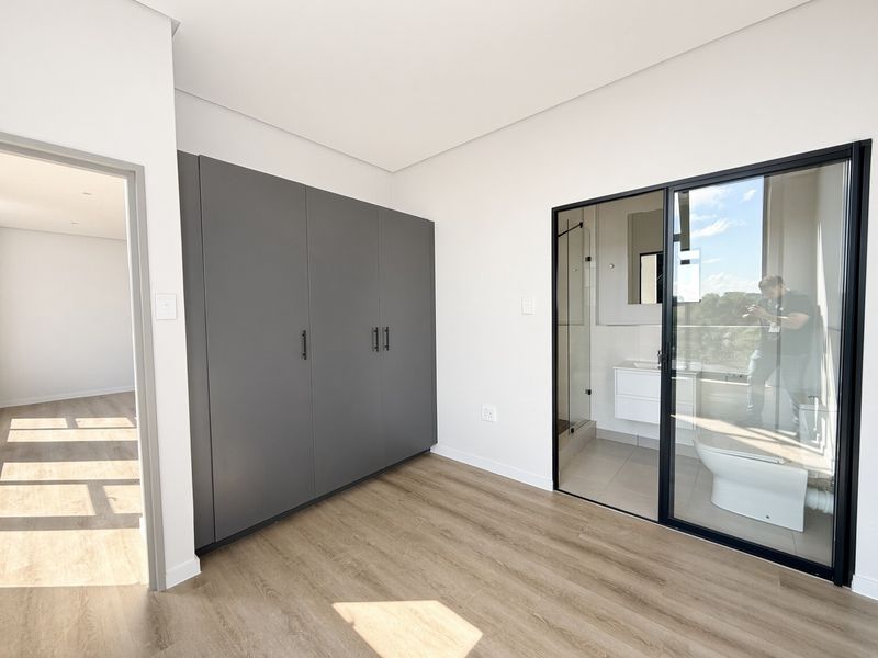 Modern bedroom featuring light laminate flooring, sleek grey built-in cupboards, and a glass-enclosed en-suite bathroom.