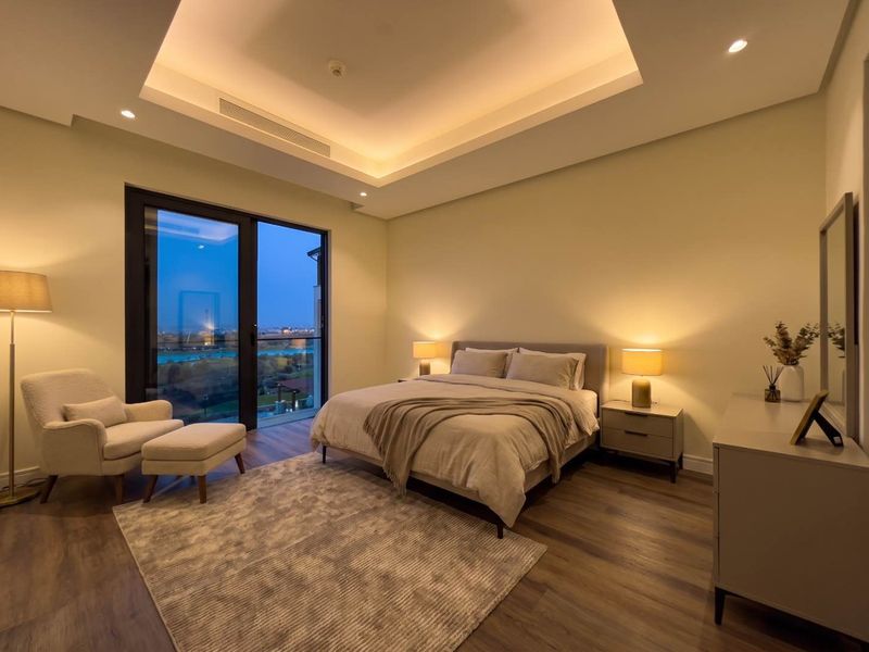 A modern, well-lit bedroom featuring a tray ceiling with recessed lighting, laminate flooring, and a sliding glass door providing access to an outdoor view.