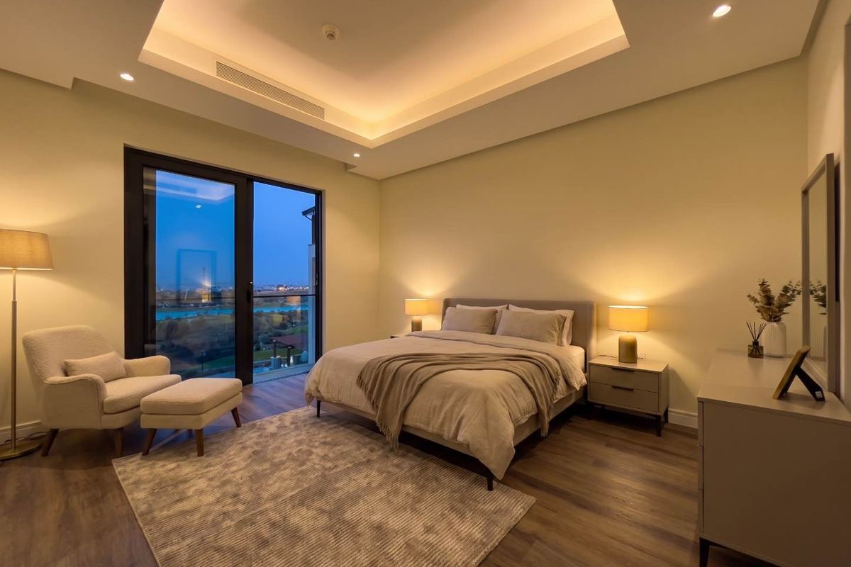 A modern, well-lit bedroom featuring a tray ceiling with recessed lighting, laminate flooring, and a sliding glass door providing access to an outdoor view.