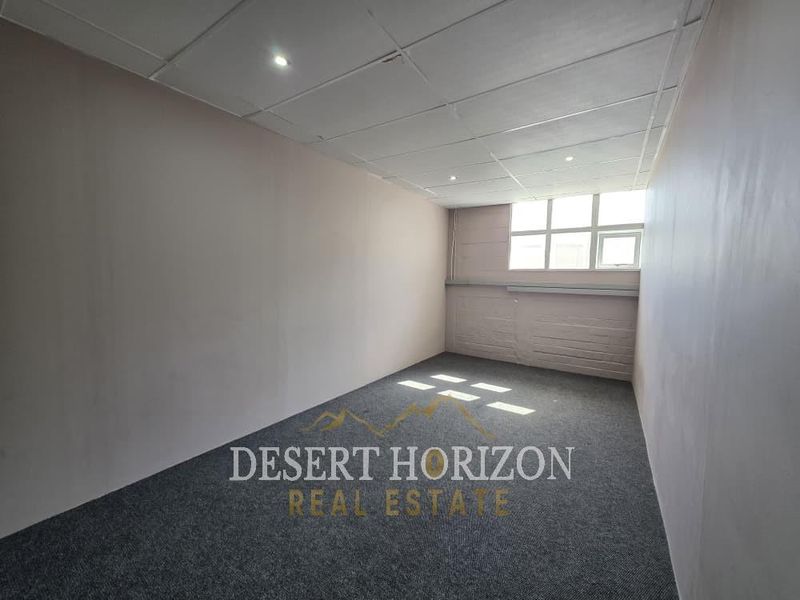 A clean, empty office space featuring grey carpet flooring, a suspended ceiling with recessed lighting, and a large window providing natural light.