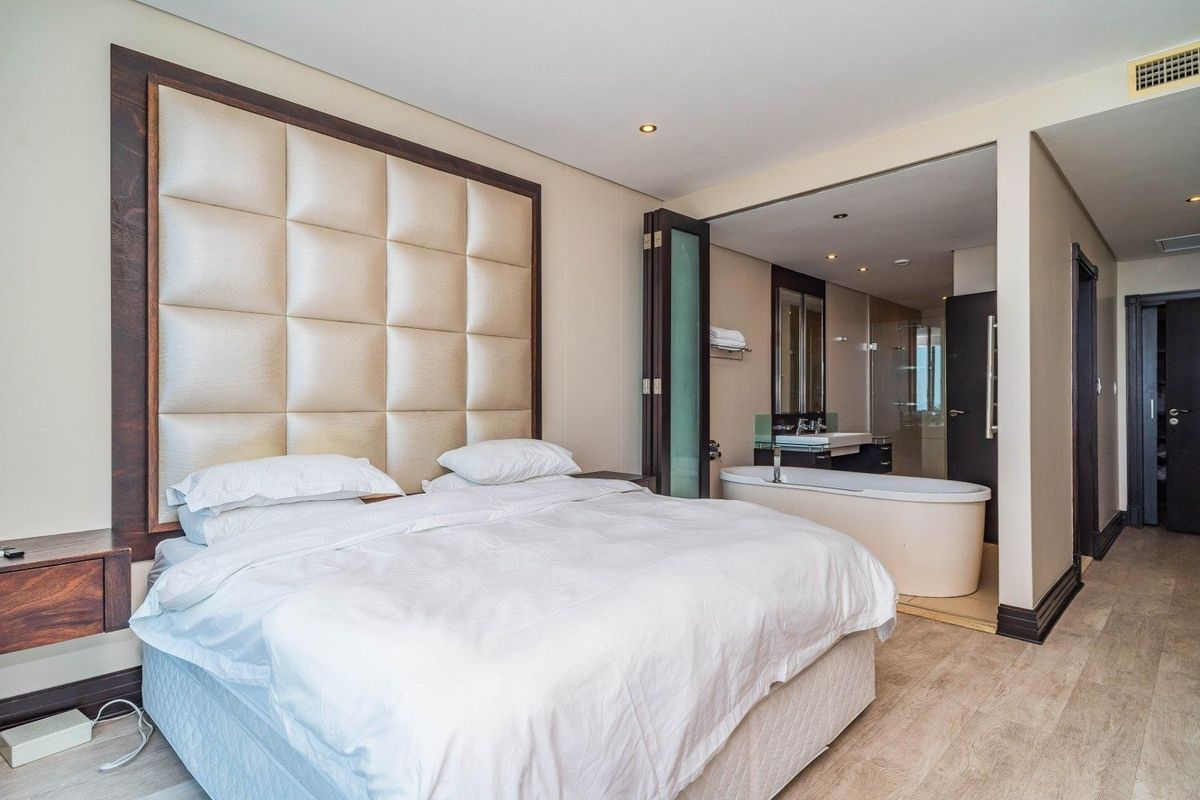 Modern bedroom featuring a large upholstered headboard, integrated open-plan bathroom with a freestanding bathtub, vanity basin, and contemporary laminate flooring.