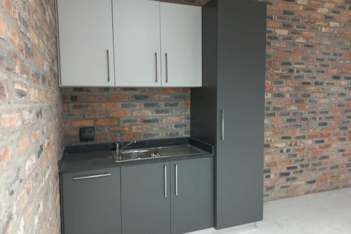 A compact kitchenette featuring modern grey cabinetry, a stainless steel sink, and a striking exposed brick wall finish.