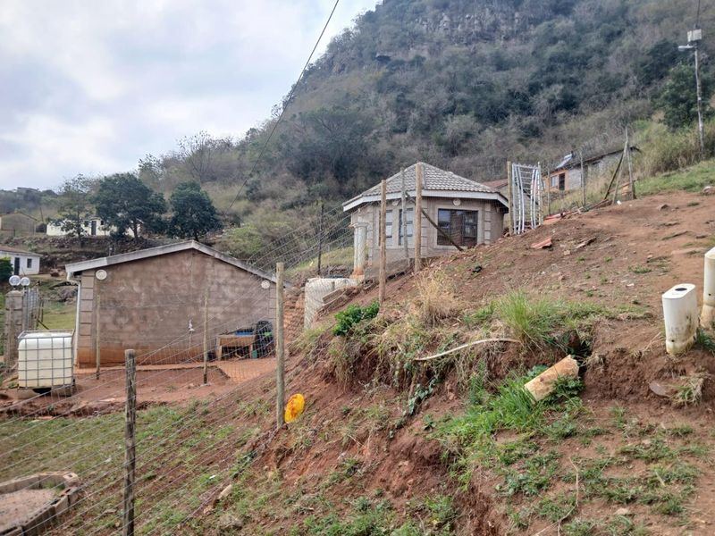 An exterior view of a property featuring concrete block structures on a sloped, undeveloped plot with wire fencing and natural hillside surroundings.