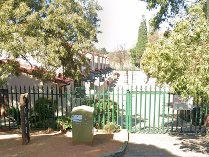 Street view of the townhouse complex entrance featuring security fencing and gated access with mature trees.