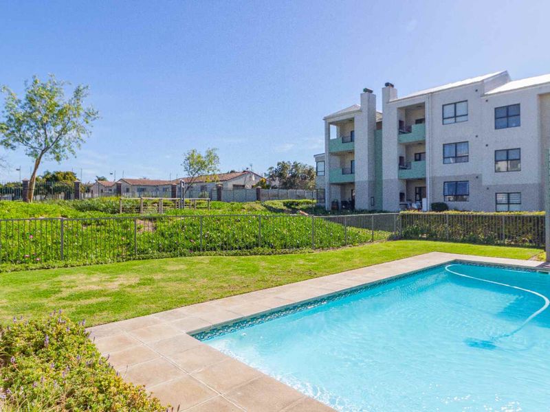 A well-maintained communal swimming pool area featuring paved surrounds, manicured lawn, and secure metal perimeter fencing within an apartment complex.