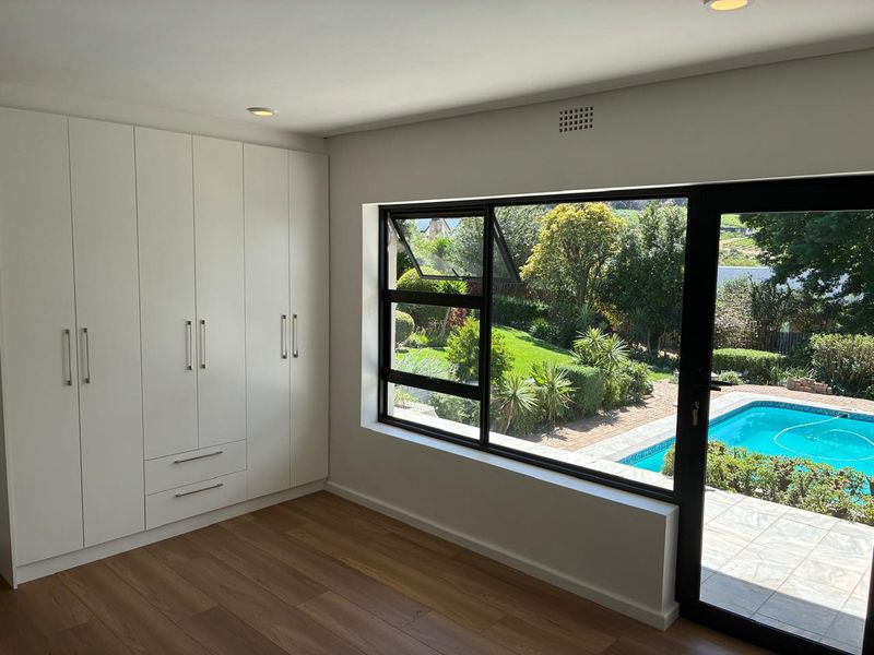 A bright, modern bedroom featuring sleek built-in cupboards, warm laminate flooring, and large windows offering a pleasant view of the garden and swimming pool.