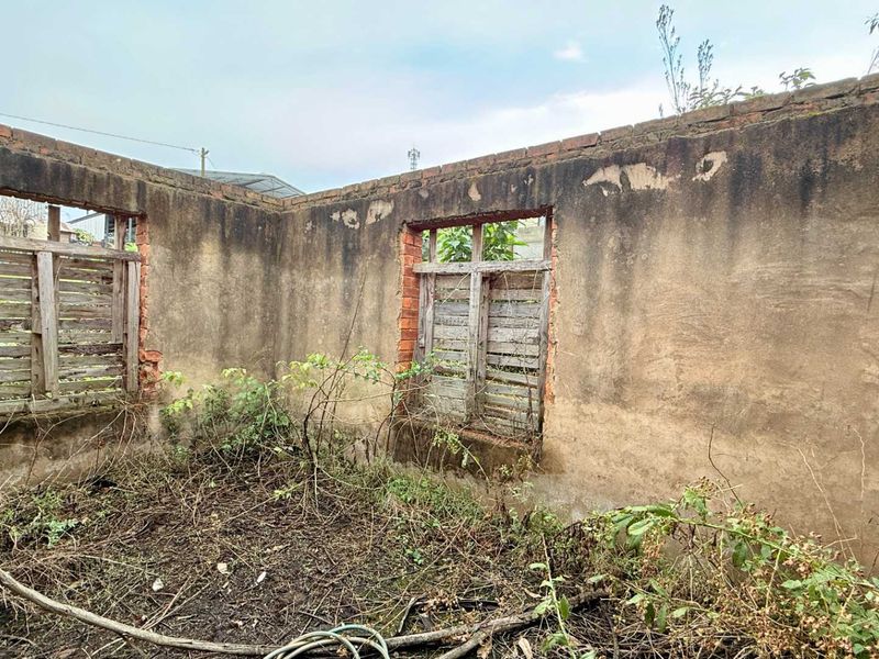 A dilapidated structure featuring weathered masonry walls and empty window frames, situated on a vacant commercial plot with significant overgrowth.