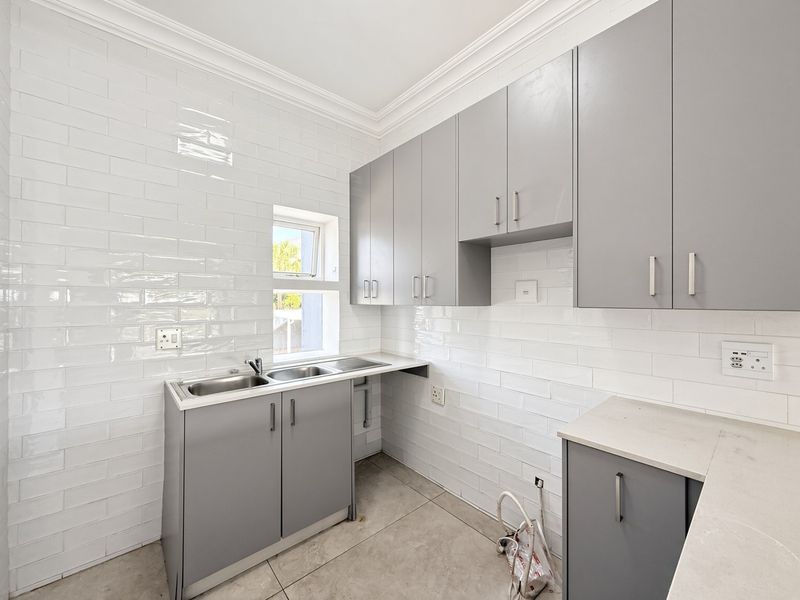 A clean, modern scullery featuring grey built-in cabinetry, stone countertops, a double stainless steel sink, and a white subway tile backsplash.