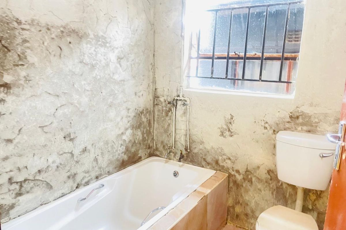 A bathroom in need of significant renovation, featuring a bathtub, toilet, and window with security bars, currently showing extensive wall surface damage.