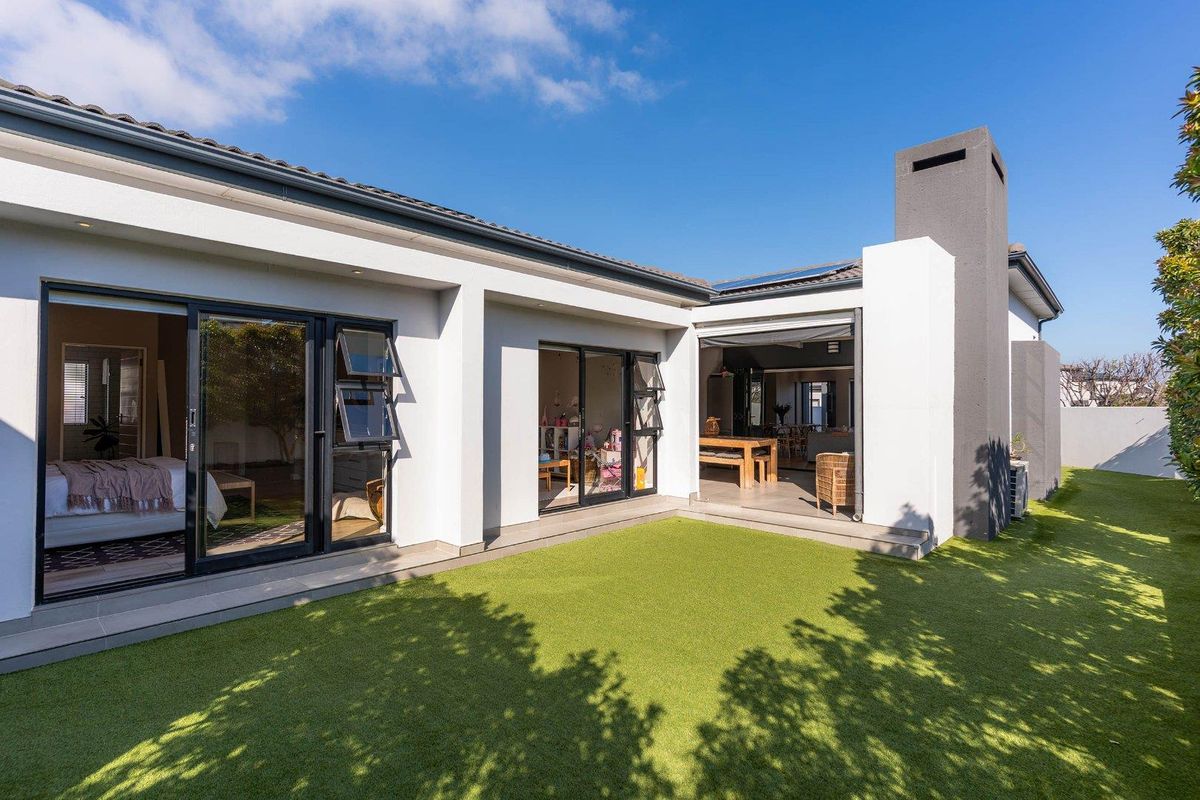 A modern rear exterior view featuring a landscaped garden, artificial lawn, and seamless indoor-outdoor flow through large sliding glass doors.