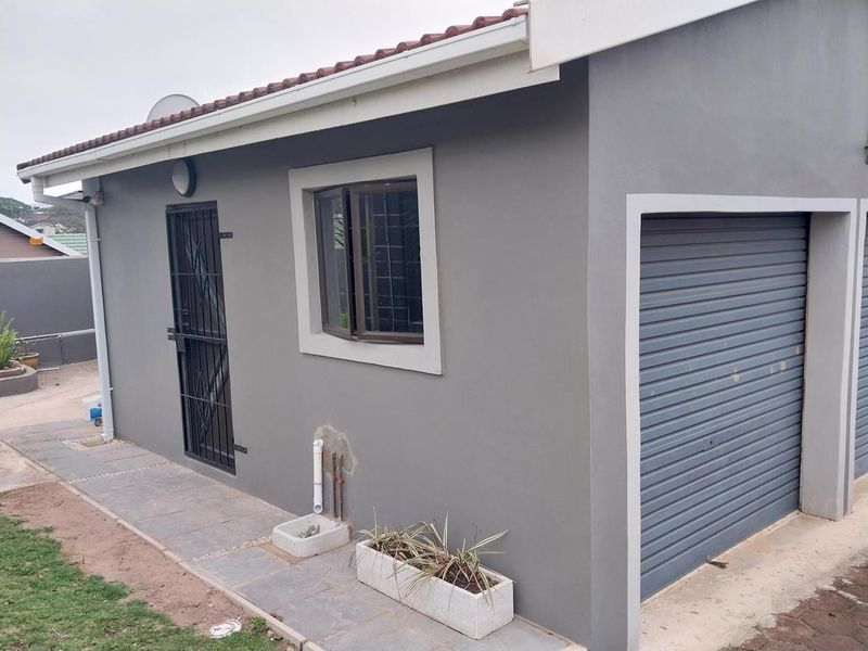 Exterior view of a cottage featuring a grey rendered facade, roller garage door, security gate, and paved walkway with visible external plumbing.