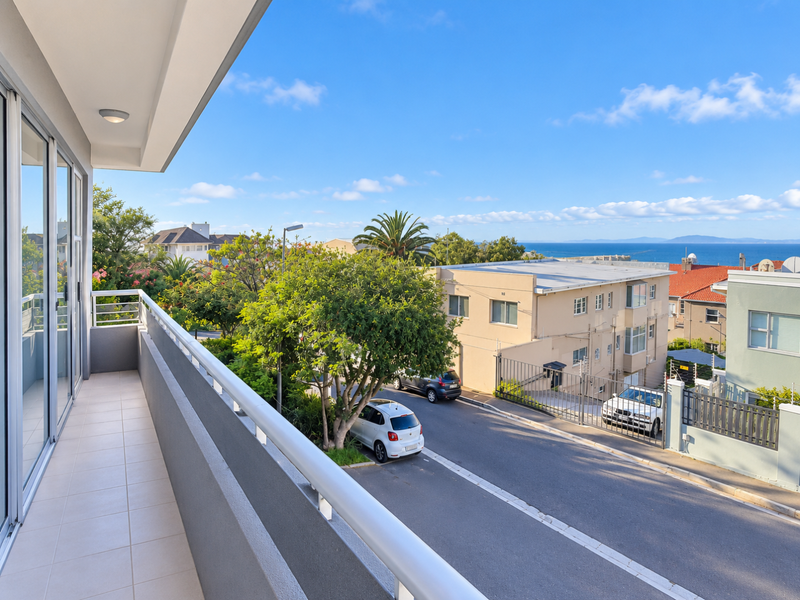 A clean, tiled balcony featuring a metal balustrade and sliding glass door access, offering an elevated view of the street and distant ocean.