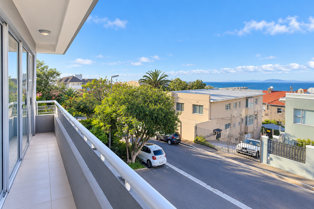 A clean, tiled balcony featuring a metal balustrade and sliding glass door access, offering an elevated view of the street and distant ocean.