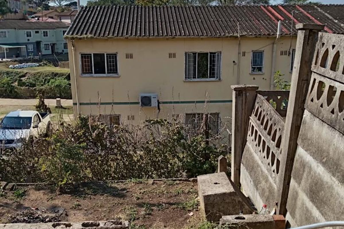 Rear exterior view of a townhouse featuring a concrete patio, boundary wall, and weathered building facade with an external air conditioning unit.