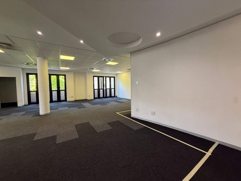 Spacious, open-plan office area featuring a suspended ceiling with recessed lighting, carpeted flooring, and multiple glass doors providing natural light.