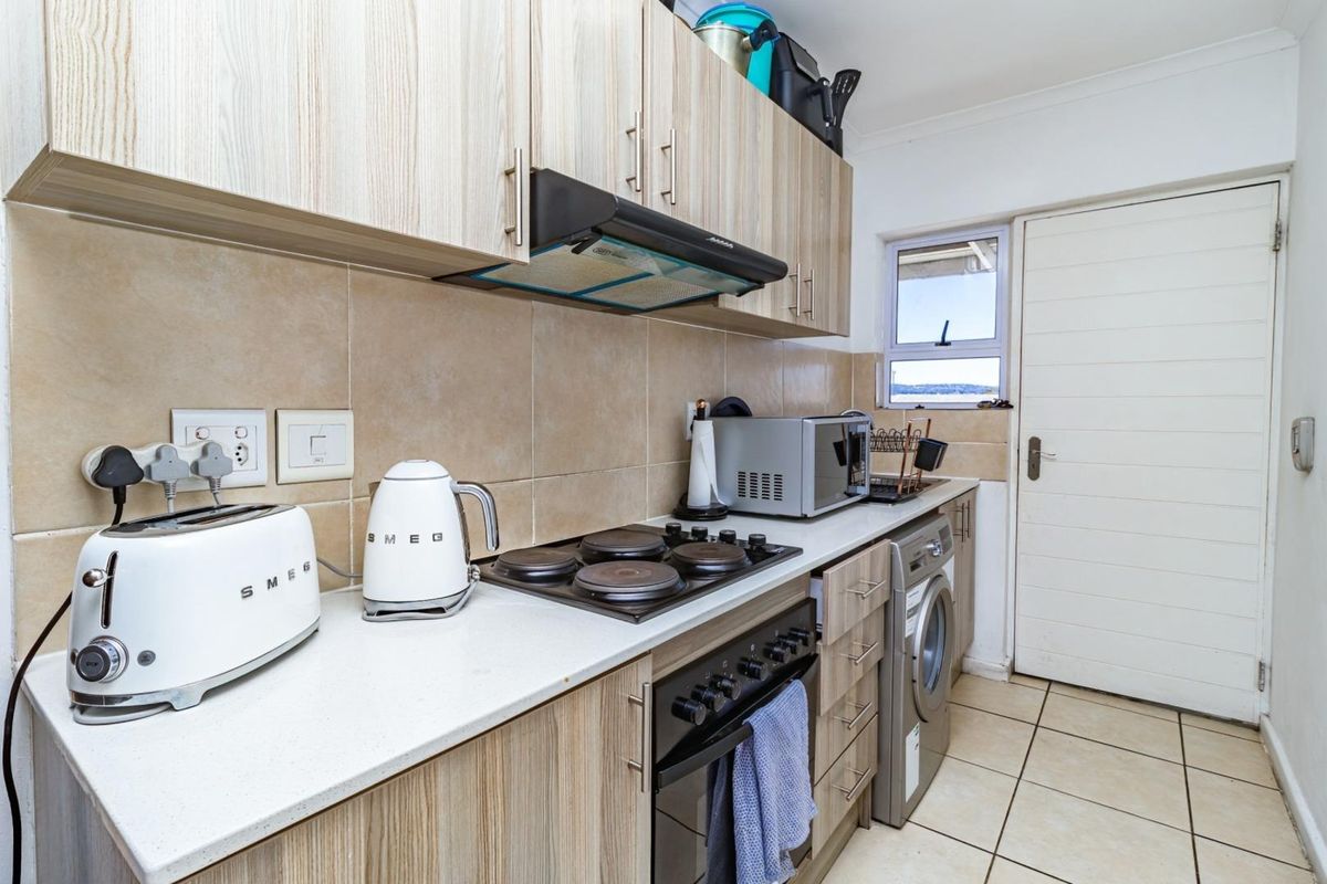 Compact kitchen featuring light wood-grain cabinetry, stone-look countertops, integrated electric hob, oven, extractor fan, and tiled flooring with direct access to an exterior door.